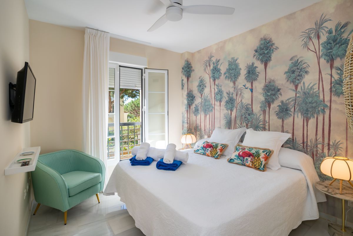 Une chambre d’hôtel avec un lit, une chaise, une télévision et une fresque représentant des palmiers.