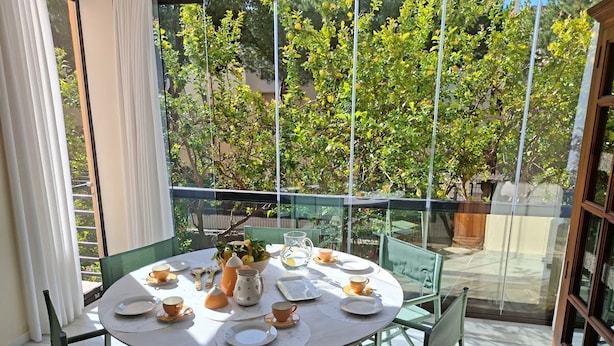 Una mesa redonda dispuesta para el té, con sillas verdes, barandilla de vidrio y vistas a árboles y edificios.