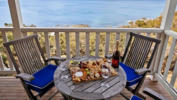 Eine Holzterrasse mit zwei Stühlen und einem runden Tisch, gedeckt mit einer Wurstplatte, Gläsern und einer Champagnerflasche.