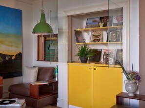 Una sala de estar con un aparador amarillo, una lámpara colgante verde, un sofá de cuero marrón y un cuadro en la pared.