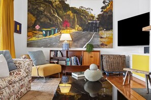 Un salon comprenant un grand tableau représentant une route, un canapé, une table basse, une bibliothèque et un téléviseur.