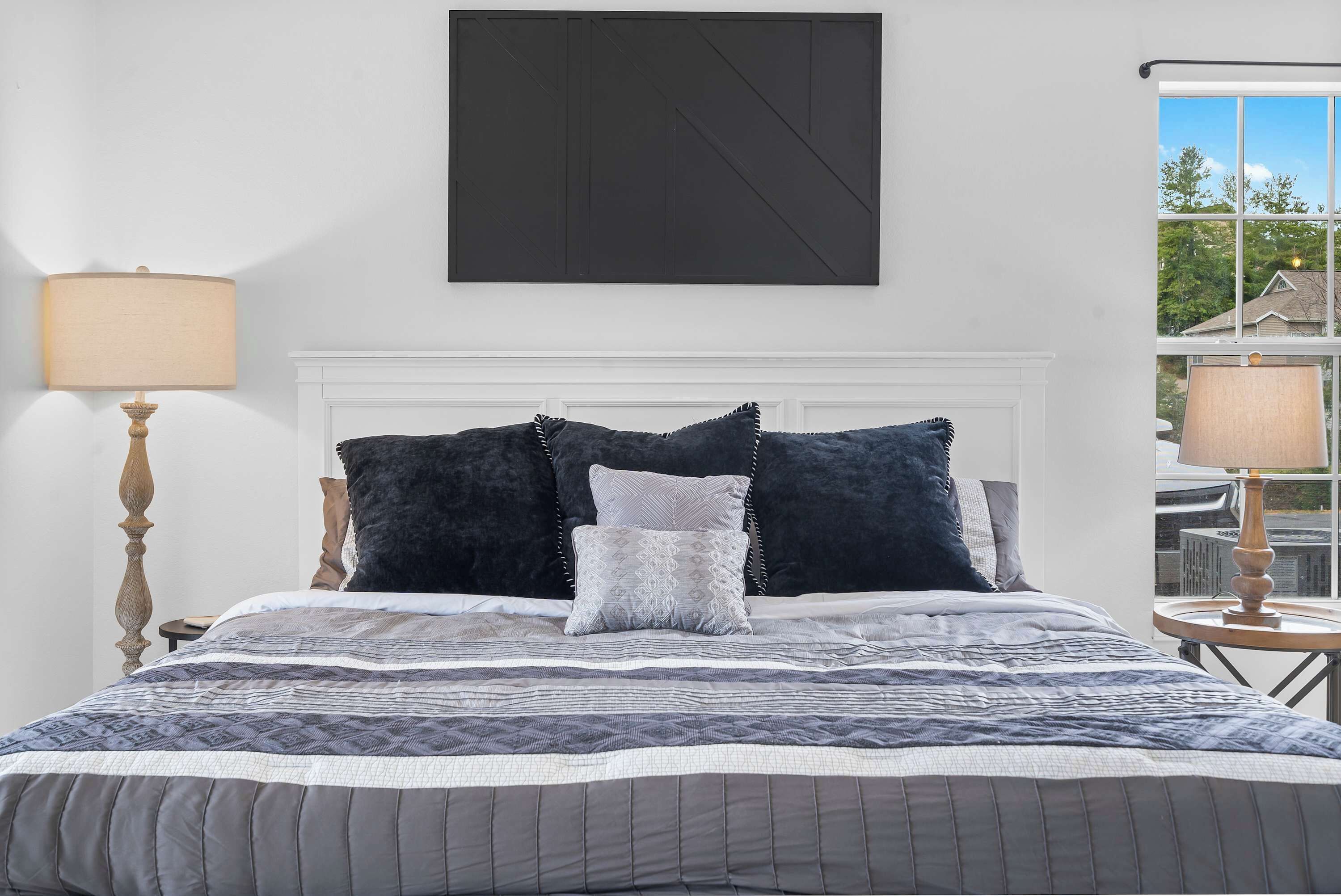 Een netjes opgemaakt bed met een geborduurd beddegoed en meerdere kussens, een bedlampje en een groot abstract schilderij aan de muur.