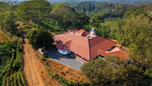 A large house with a red roof, surrounded by lush greenery and a hilly landscape.