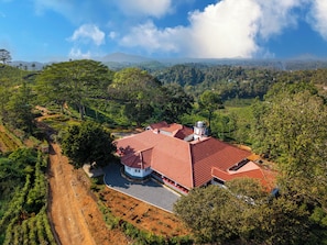 A large house with a red roof, surrounded by lush greenery and a hilly landscape.