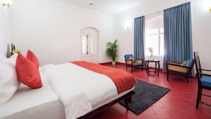 A hotel room with a bed, a desk, a chair, a potted plant, and a window with curtains.