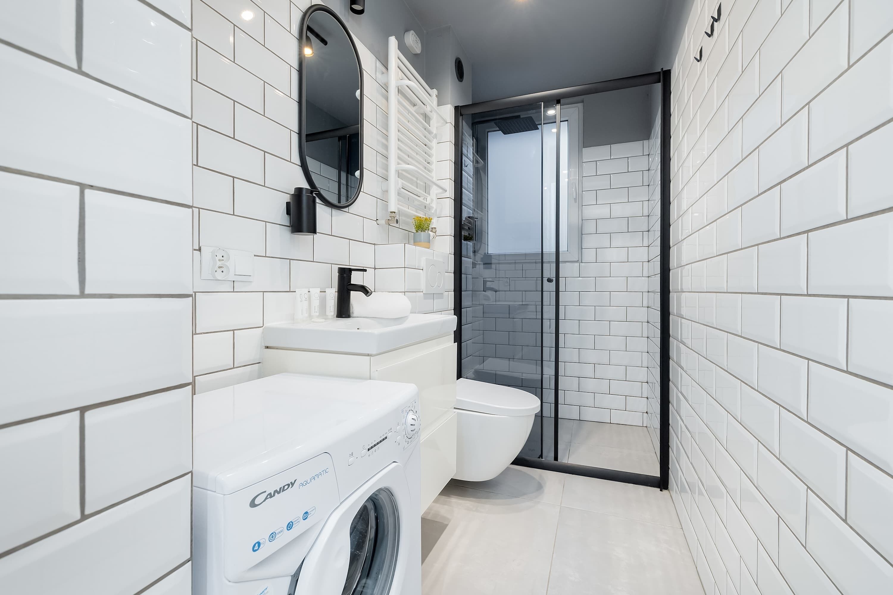 A modern bathroom with white tiles, a washing machine, a toilet, and a glass shower enclosure.