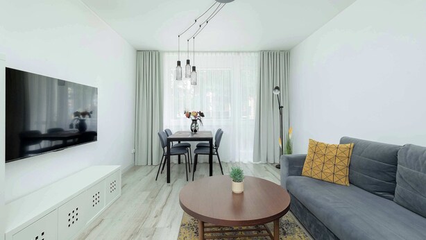 A modern living room with a flat-screen TV, a grey sofa with a patterned cushion, a round wooden coffee table, and a dining area with a table and chairs.