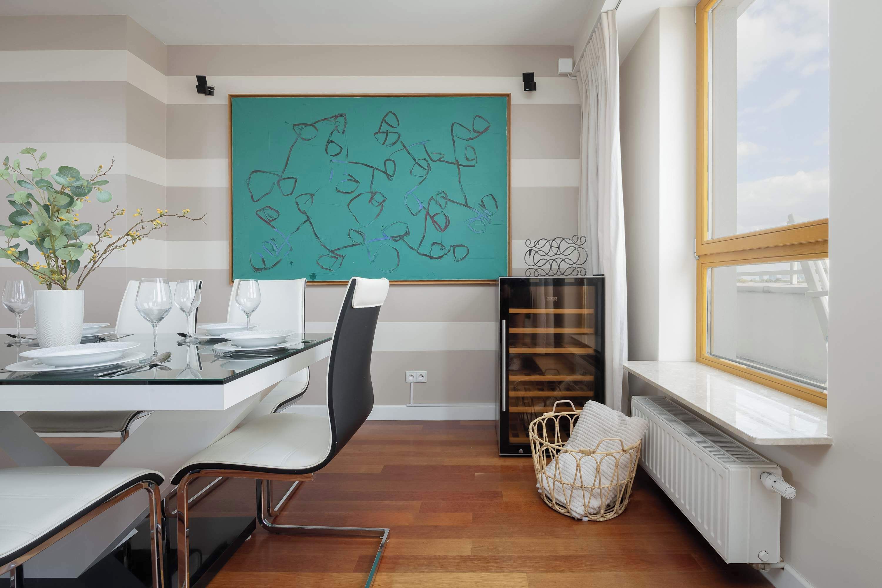 Un comedor moderno con una mesa blanca dispuesta para cuatro, una silla negra, un radiador blanco y un cuadro abstracto grande en la pared.
