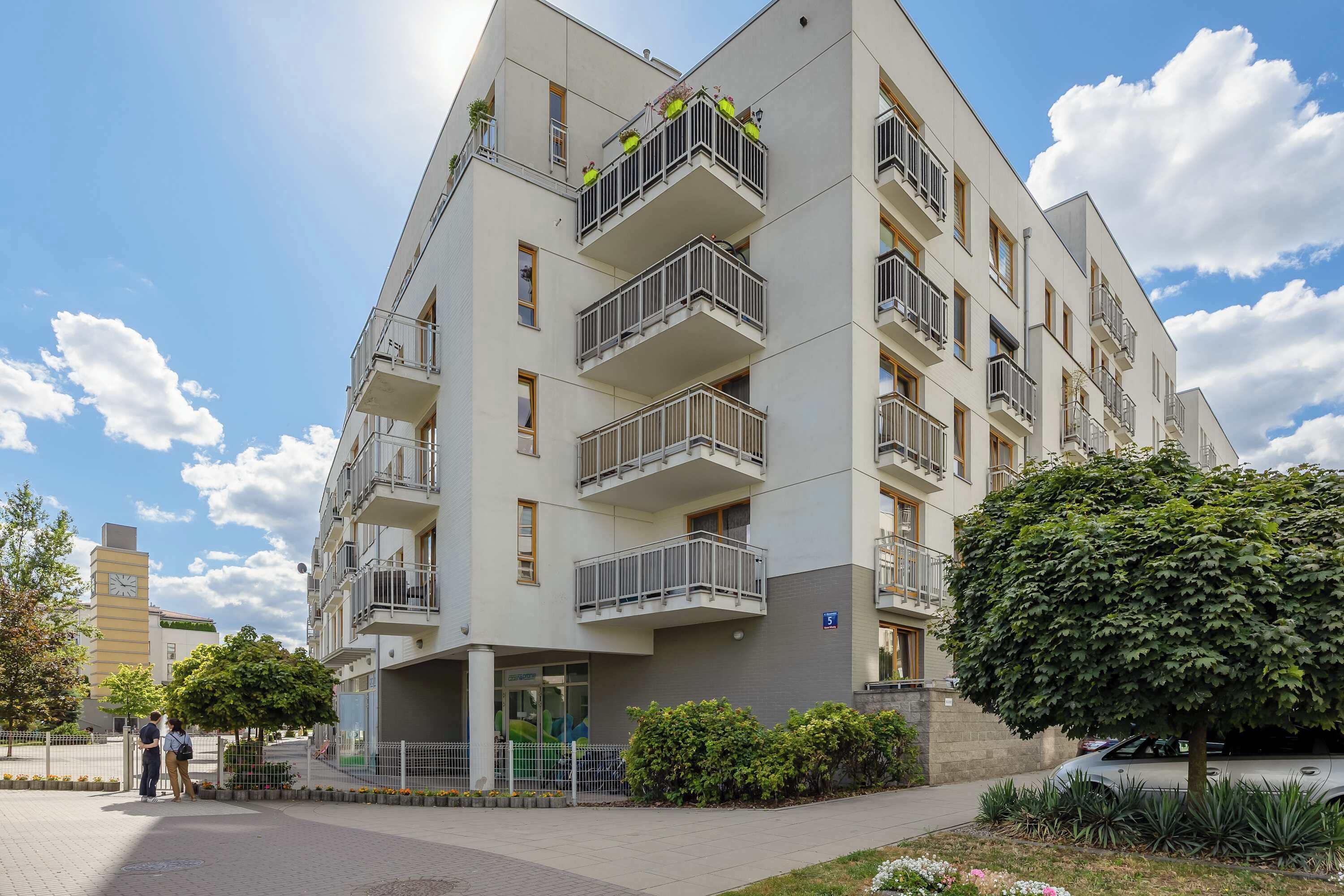 Un edificio residencial de varios pisos con balcones, entrada protegida y zona de estacionamiento.