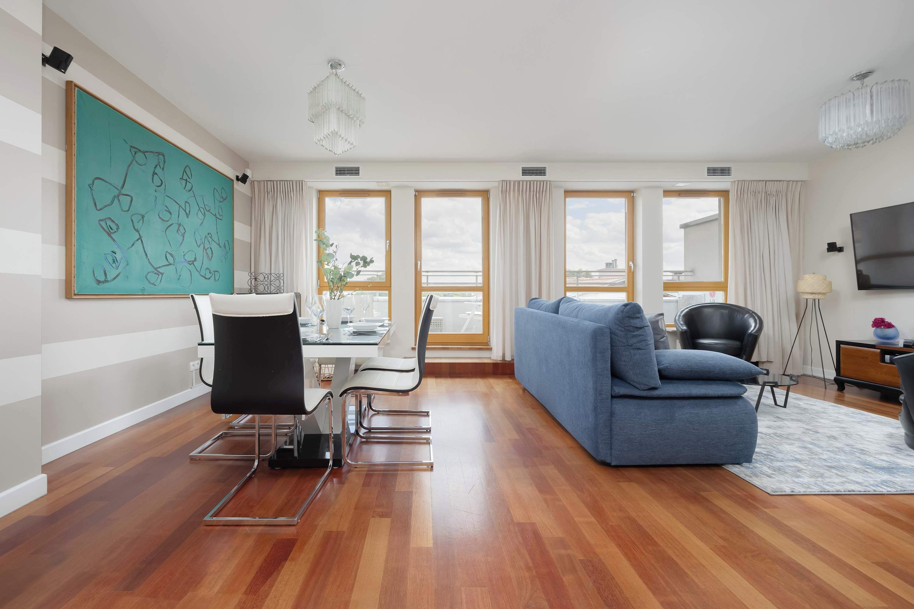 A modern living room with a blue sofa, dining table, and chairs. There are large windows with curtains, a chandelier, and a flat-screen TV.