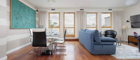 A modern living room with a blue sofa, dining table, and chairs. There are large windows with curtains, a chandelier, and a flat-screen TV.