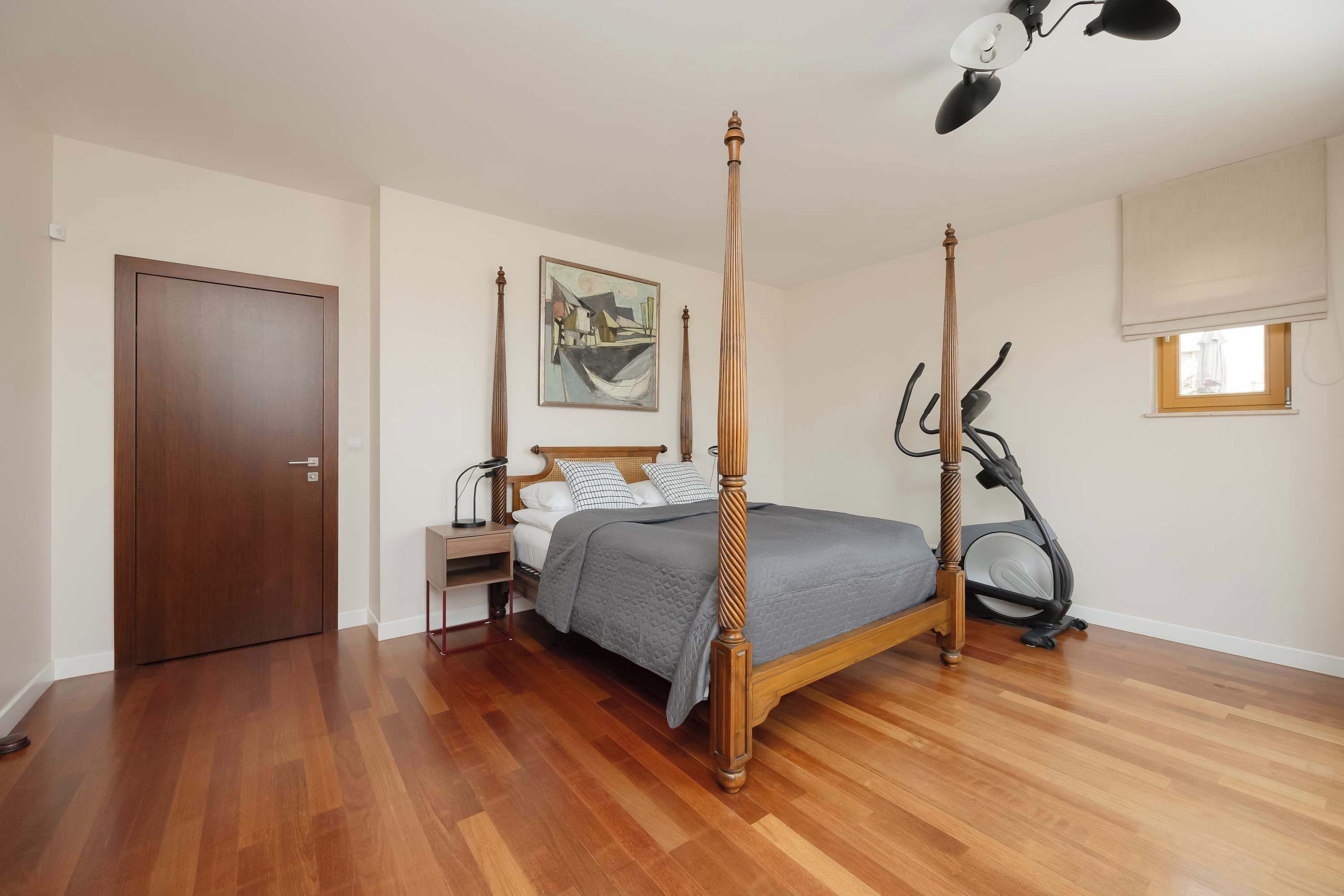 A bedroom with a four-poster bed, a standing lamp, a painting on the wall, and a window with a blind.