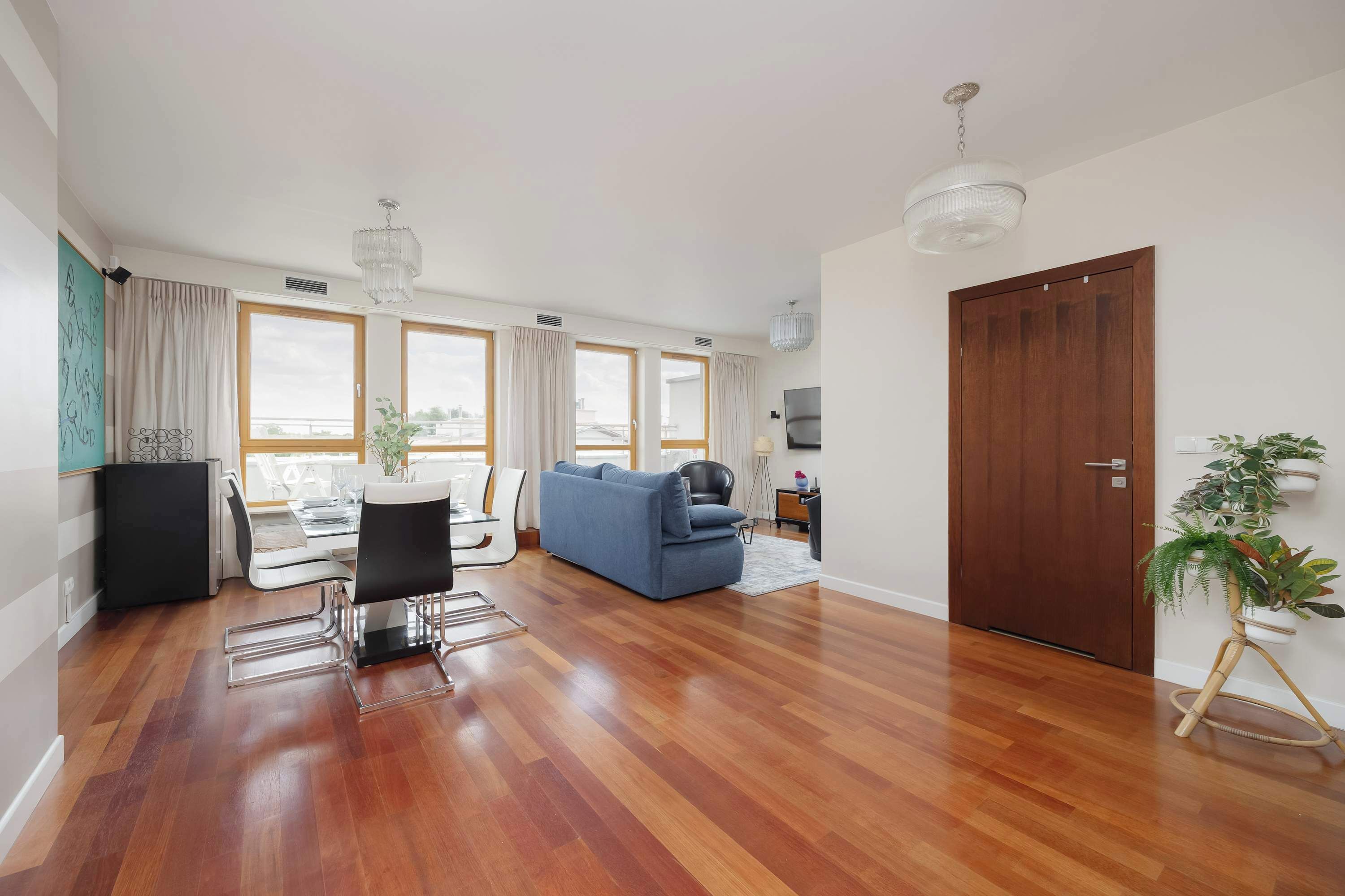 Un salon moderne comprenant un coin repas, un parquet, un canapé bleu, une table à manger avec des chaises et une grande fenêtre avec des rideaux.