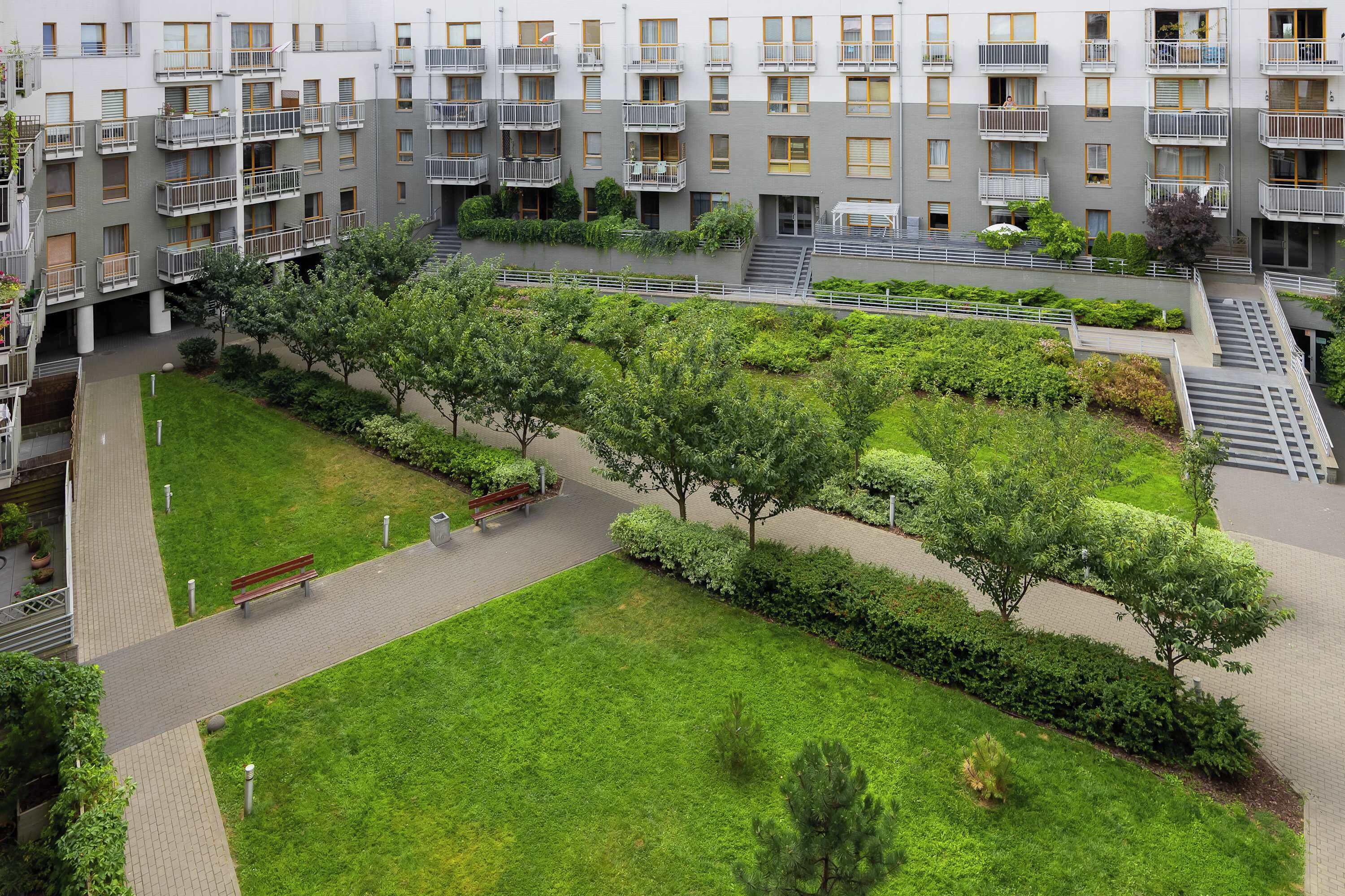 A multi-story residential building with balconies, a central courtyard with a pathway, benches, and greenery.