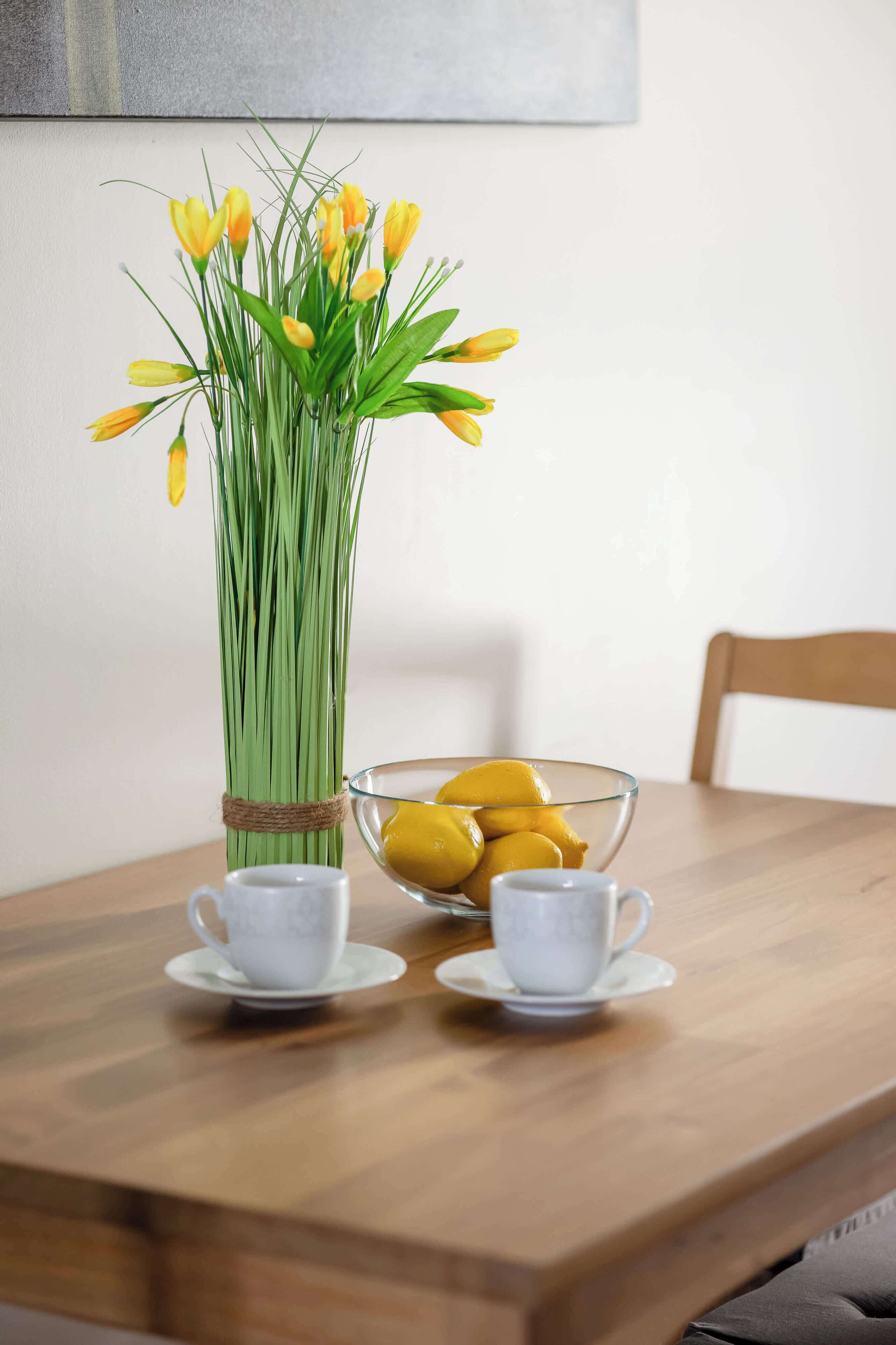 A wooden table with a vase of yellow tulips, a glass bowl of lemons, and two white cups on saucers.