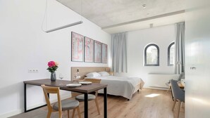A modern hotel room with a bed, a desk, a chair, a dining table, and a vase with flowers.
