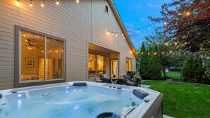 A hot tub in front of a house with a patio area, string lights, and a lawn.