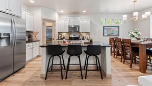 A modern kitchen with white cabinets, stainless steel appliances, a central island with bar stools, and a dining area with wooden chairs and a chandelier.