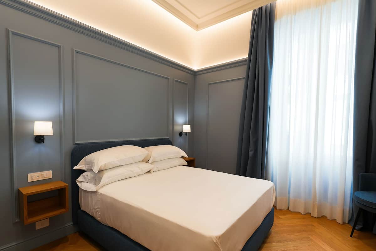 Un lit bien fait, avec du linge de lit blanc, une table de chevet en bois sur laquelle se trouve une lampe, et une chaise près de la fenêtre, devant des rideaux bleus.