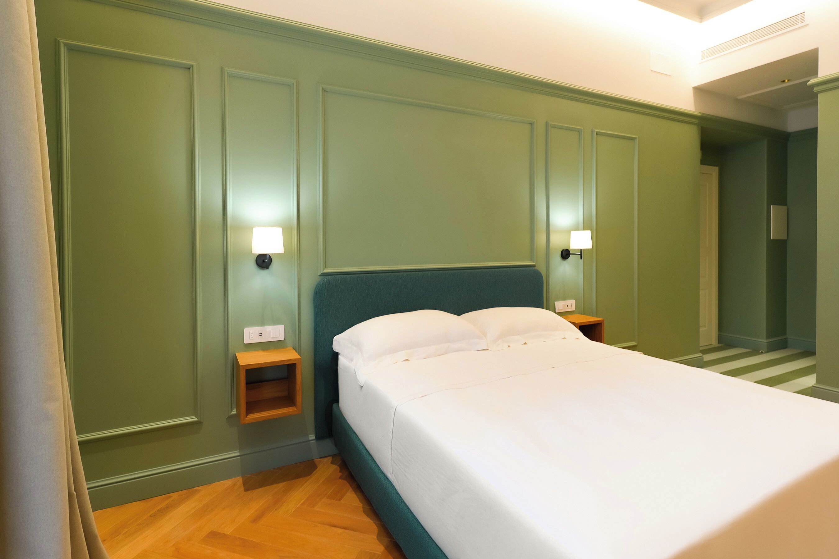 Cama bem arrumada em quarto com paredes verdes e piso de madeira. Há duas mesas de cabeceira com luminárias.