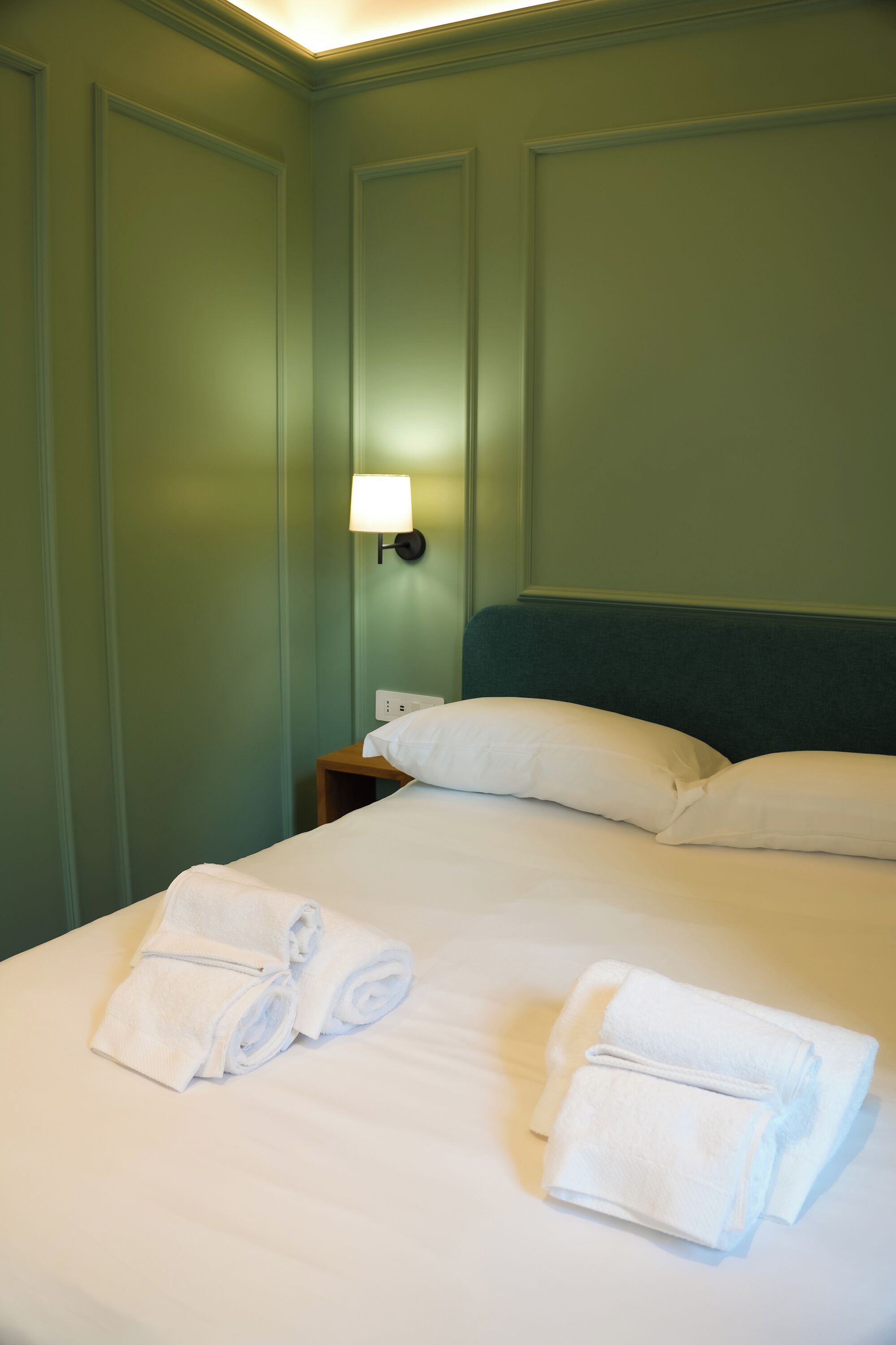 Un lit bien fait, avec du linge de lit et des serviettes blanches, une lampe de chevet et un mur vert.