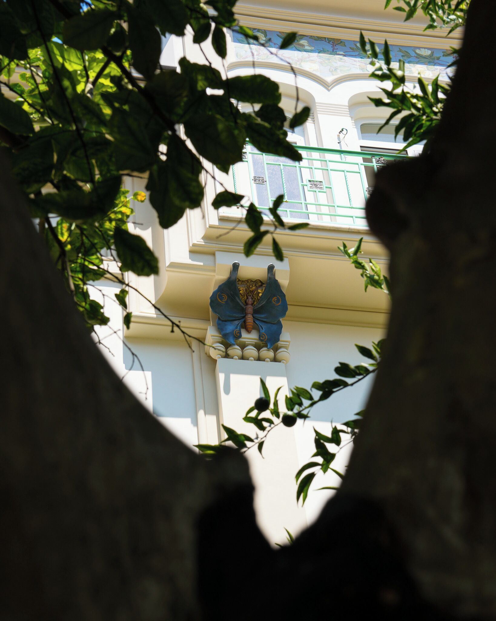 Vista a través de una rama de árbol, enfocada en la fachada de un edificio con una escultura decorativa.