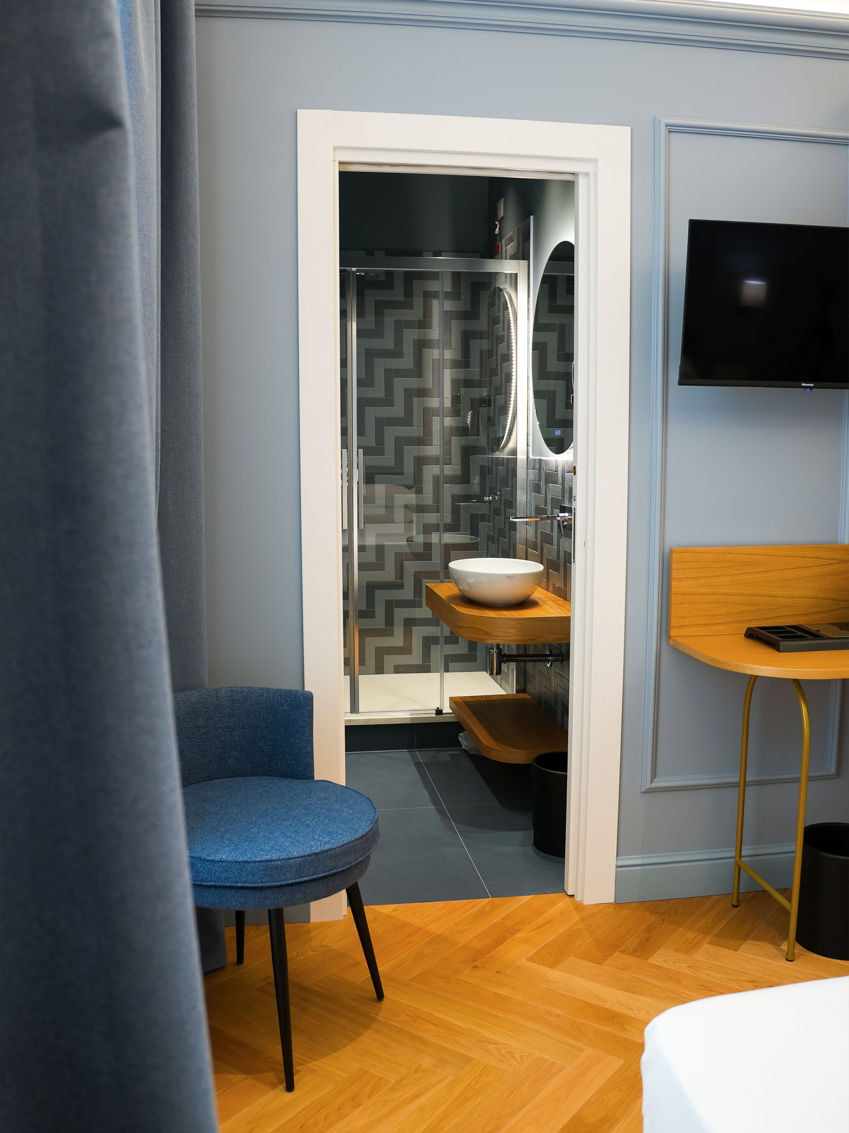 Un baño moderno con ducha, lavamanos y espejo. Hay una silla azul y una mesa de madera con un teléfono.