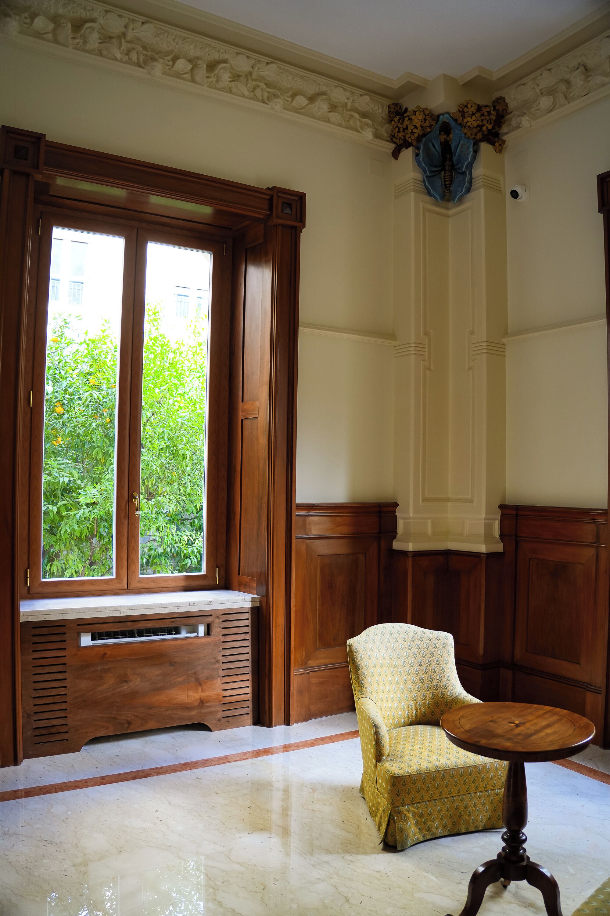 Een kamer met een groot raam, een houten bankje, een gestreept fauteuil en een ronde houten tafel.