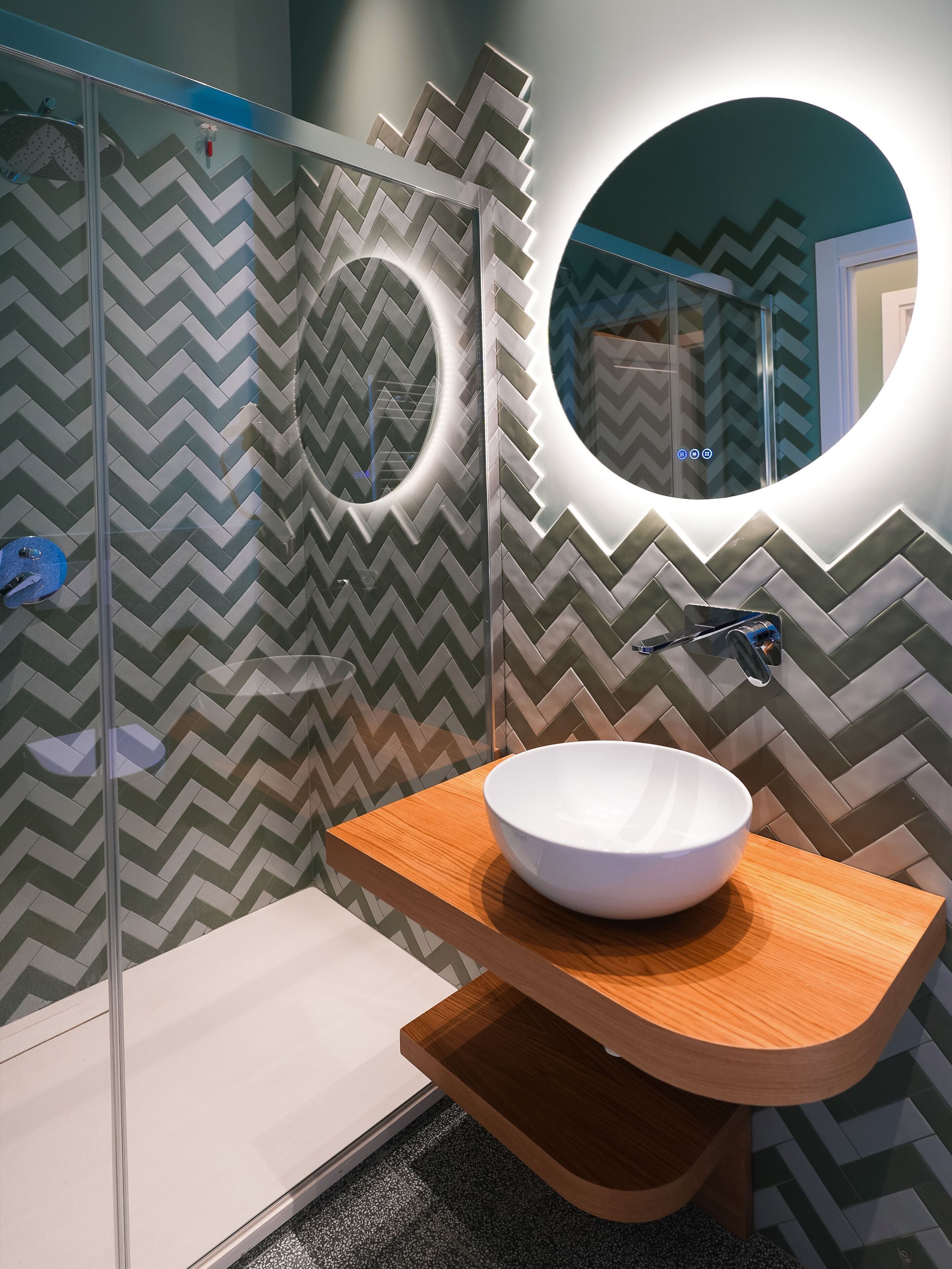 A modern bathroom with a zigzag patterned wall, a round mirror, and a white vessel sink on a wooden countertop.