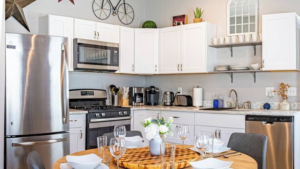 A modern kitchen with white cabinets, stainless steel appliances, and a dining area with a wooden table set for four. - Stylish 2BR/2BA House in The Grove w/ Free Parking (St. Louis)