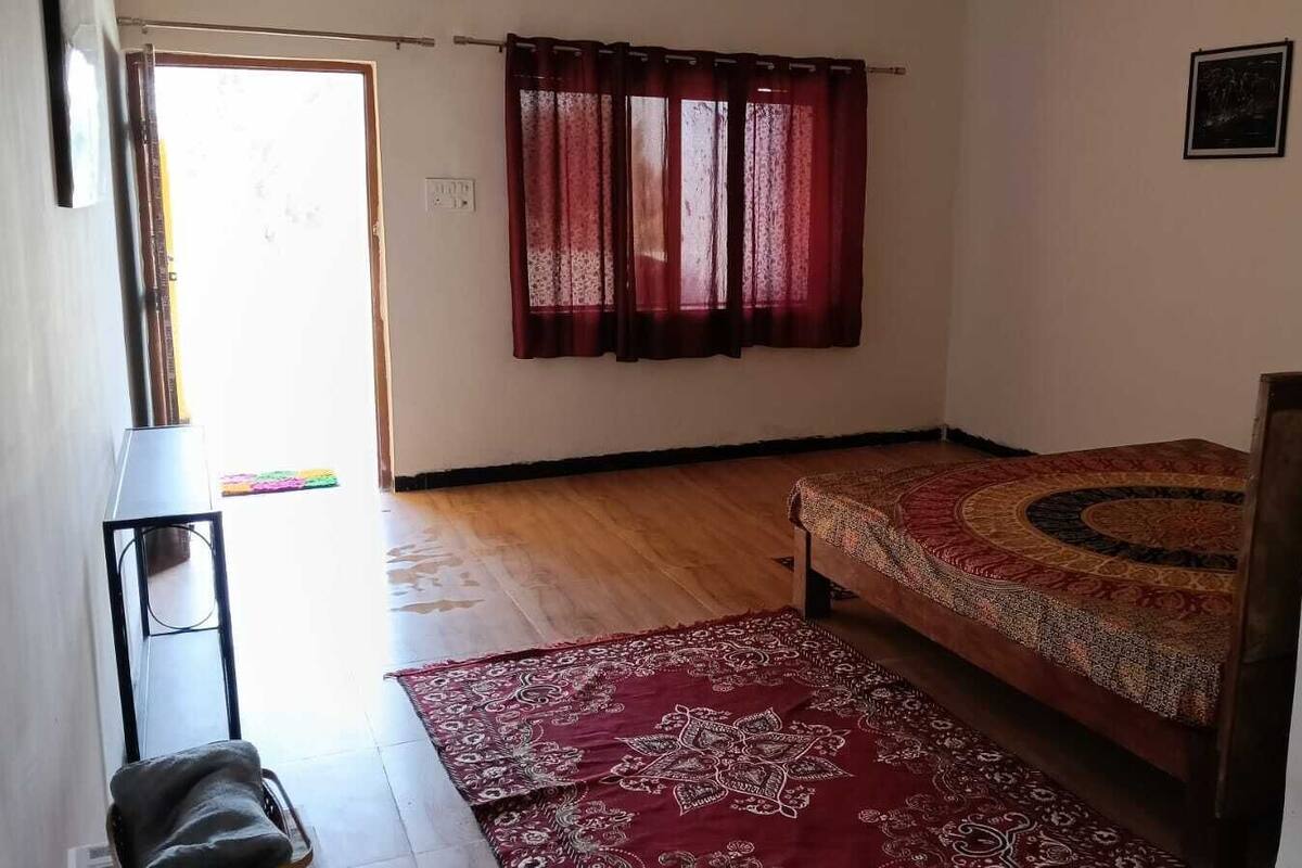 Un dormitorio con suelo de madera, una cama con colcha estampada, una alfombra roja, una mesita y una ventana con cortinas.
