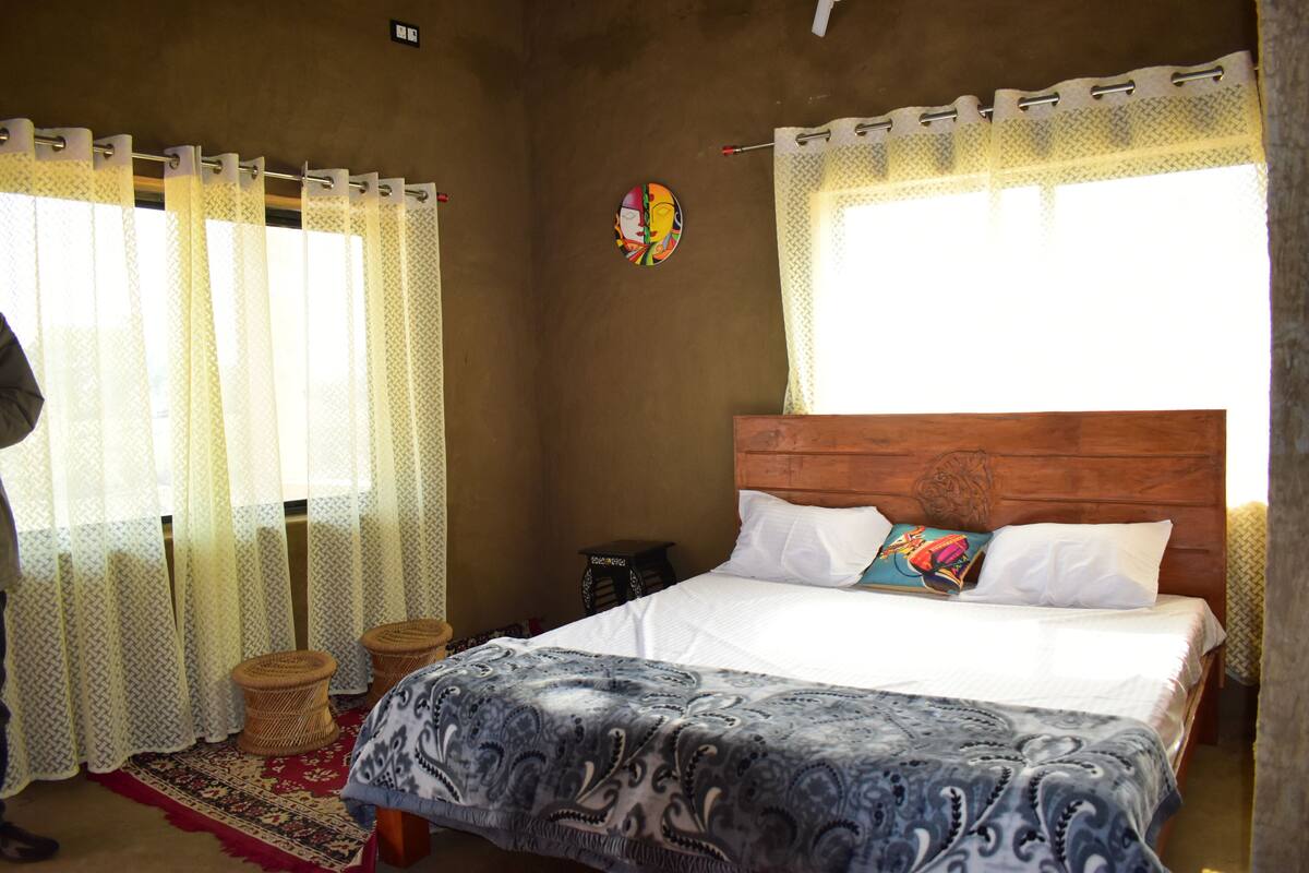 Un dormitorio con cabecera de madera, cama con ropa de cama blanca y estampada, dos taburetes de fibras tejidas, una alfombra estampada en rojo y negro, y cortinas de encaje.