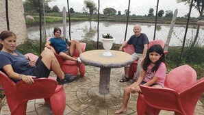 A group of people seated around a round table with a central vase, outdoors with a view of a water body and greenery in the background. - Ajinkya Jungle lake farms resort (Chimur)