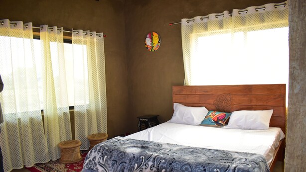 A bedroom with a wooden headboard, a patterned bedspread, and a red and white patterned rug. There are two woven stools and a small table with a lamp. - Ajinkya Jungle lake farms resort (Chimur)