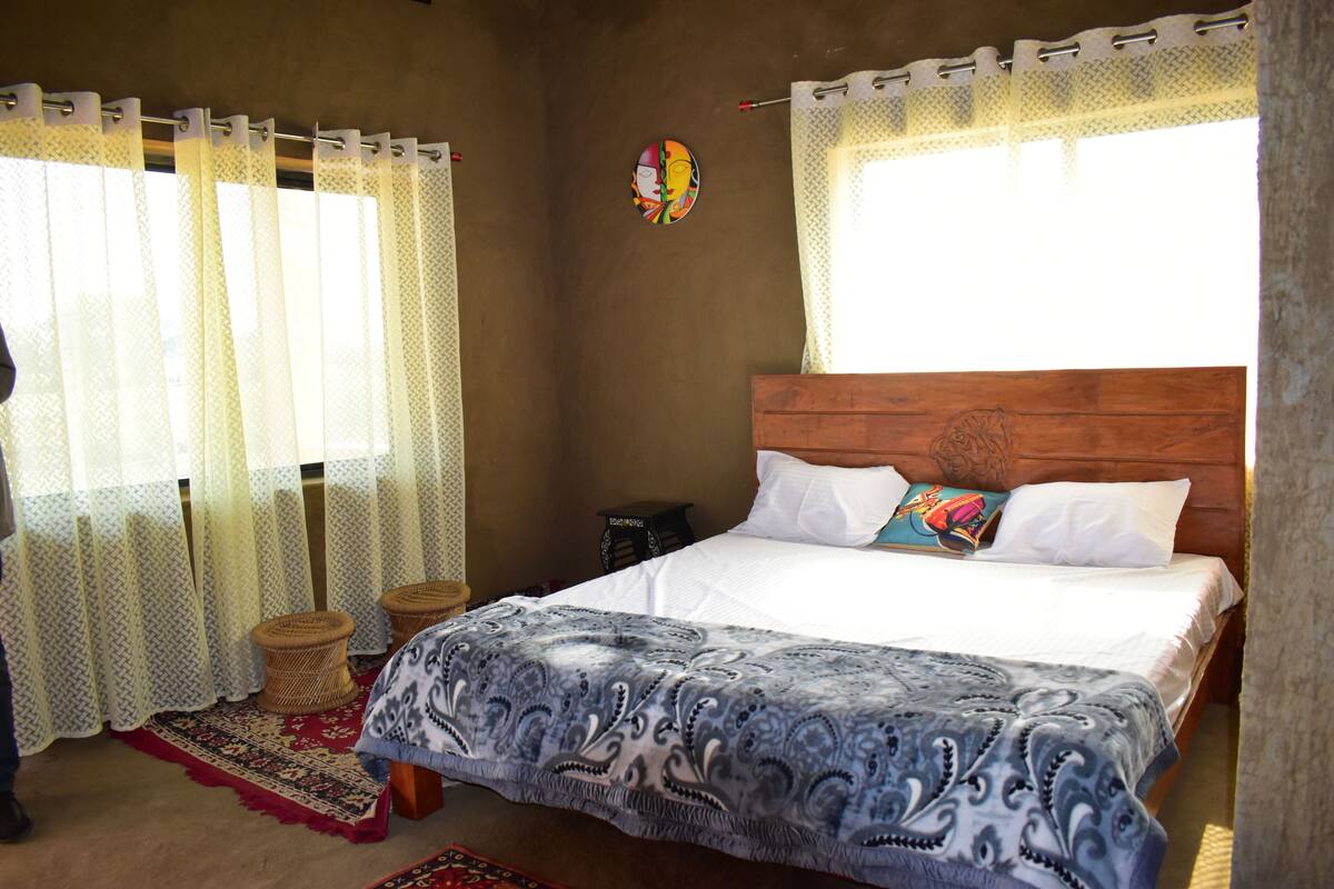 Un dormitorio con cabecera de madera, colcha estampada y alfombra roja y blanca con diseño. Hay dos taburetes tejidos y una pequeña mesa con una lámpara.