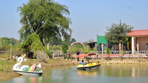 A calm river with two boats, one shaped like a swan and another with a red canopy. There are trees and a building with a green roof in the background. - Ajinkya Jungle lake farms resort (Chimur)