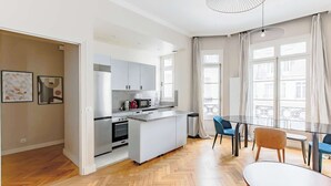 A modern kitchen with a central island, a dining area with a glass table, and large windows with curtains. - Elegant apartment with suite - Arc de triomphe (Paris)