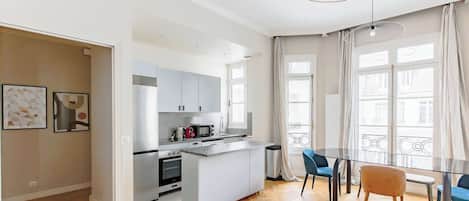 Une cuisine moderne dotée d’un îlot central, d’un coin repas avec une table en verre et de grandes fenêtres avec des rideaux.