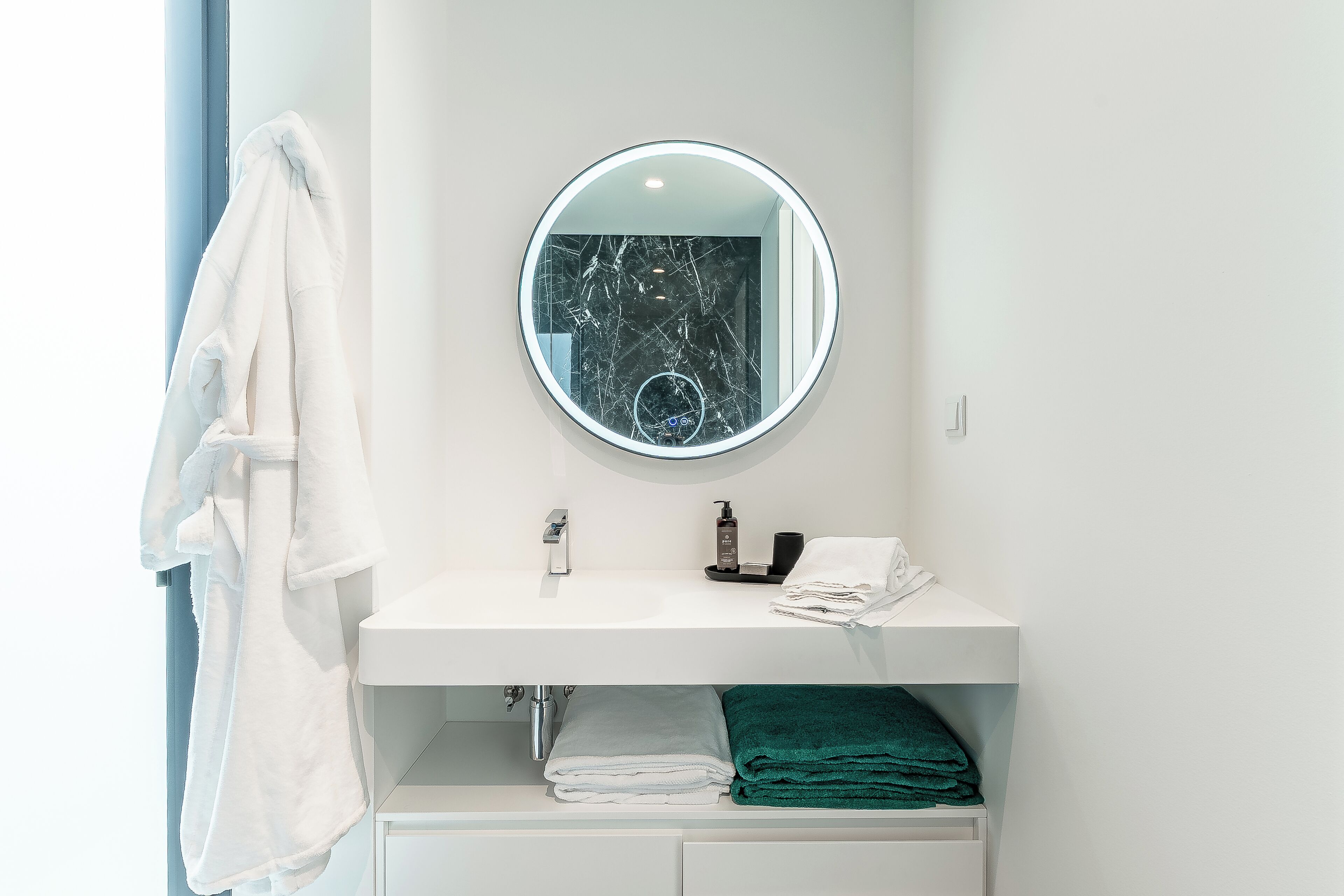 A bathroom with a white sink, a round mirror, a towel rack with a white towel, and a green towel on the counter.