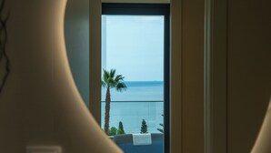 Ein Zimmer mit Meerblick durch ein großes Fenster, eine Palme und eine kleine Sitzecke mit einem Handtuch.