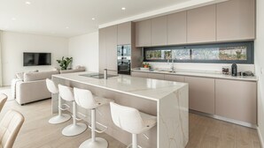 Eine moderne Küche mit einer zentralen Kochinsel, Barhocker, einem eingebauten Backofen und einem großen Fenster mit Blick auf einen Garten.