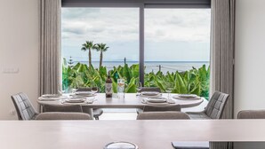 Ein Essbereich mit einem Tisch für vier Personen, großen Fenstern mit Vorhängen und Blick auf das Meer und viel Grün.