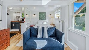 A bedroom with a bed, a blue sofa, a nightstand, a lamp, and a view of the outdoors. - Little House at Saskadena, right at the base of Ski Hill (Pomfret)