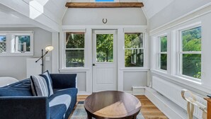 A room with a high ceiling, white walls, a dark blue sofa, a round wooden table, and large windows offering a view of greenery. - Little House at Saskadena, right at the base of Ski Hill (Pomfret)