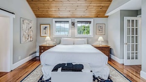 A bedroom with a wooden ceiling, a large bed, two windows, and a door. - Saskadena Lodge & Little House, Full Property (Pomfret)