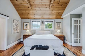 A bedroom with a wooden ceiling, a large bed, two windows, and a door.