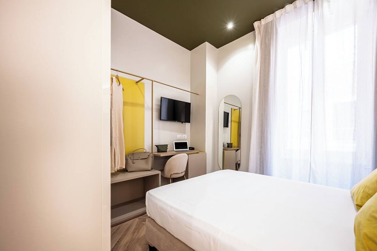 Habitación de hotel con cama, escritorio con una computadora portátil, silla, televisor montado en la pared y espejo.