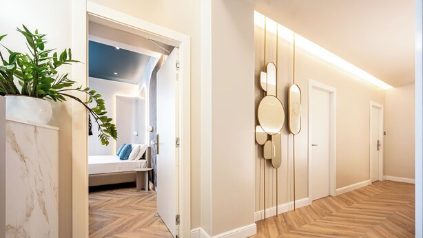 Un couloir avec un parquet en chevrons, une plante en pot, un lit avec des oreillers et un miroir fixé au mur.