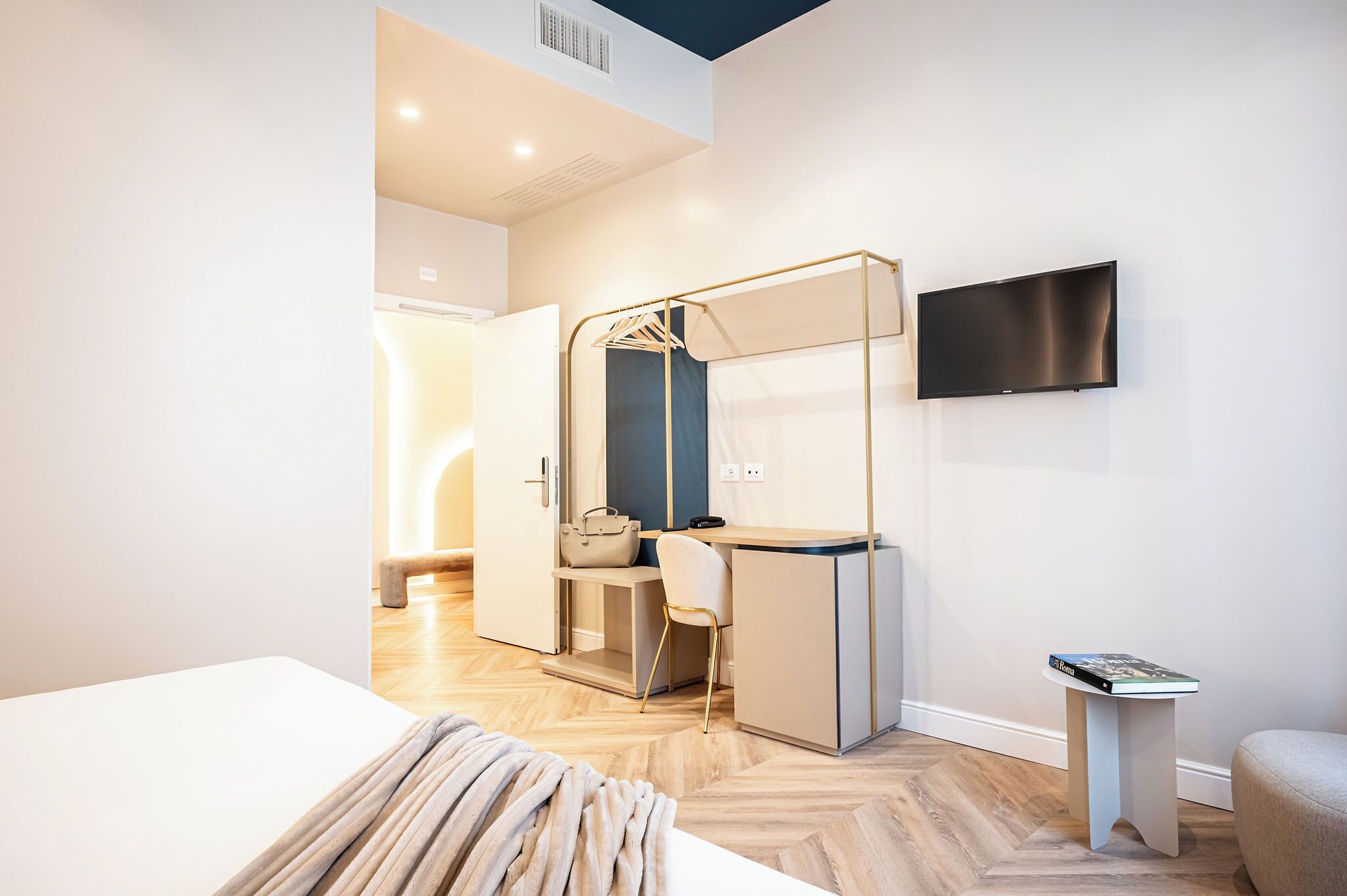 A modern hotel room with a bed, a desk with a chair, a TV mounted on the wall, and a small table with a book.