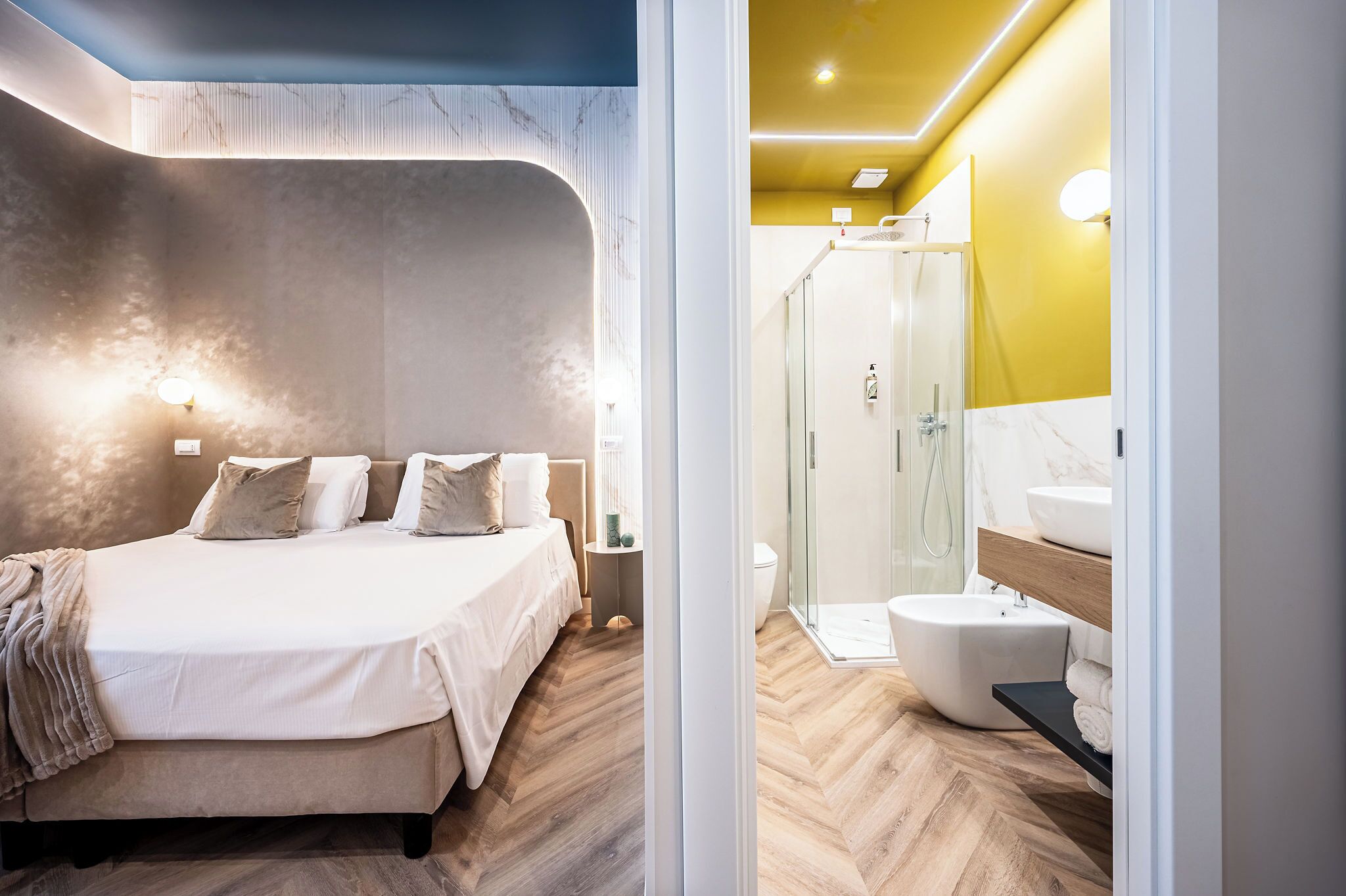 Un dormitorio moderno con una cama grande, mesitas de noche y una mesita auxiliar. Junto a él, un baño con ducha de vidrio, lavabo e inodoro.